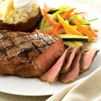 Thinly sliced, medium rare New York steak, grilled with criss cross pattern, on plate with baked potato topped with butter and sour cream and julienne vegetables