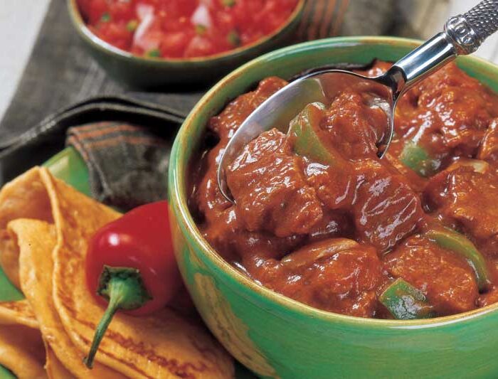 tritip_recipes Green plate with green bowl of chile with meat chunks and green pepper smothered in gravy. Red pepper garnish and folded tortilla on edge of plate. Small bowl of salsa in background