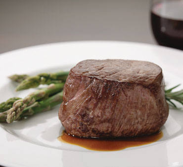 white plate with baseball cut of cooked beef and asparagus. Hint of red wine glass in background