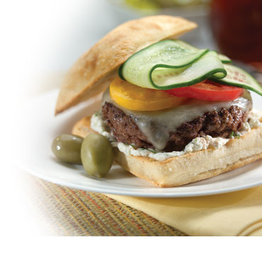 Burger on toasted sourdough bun and bed of creamy cheese, topped with melted jack, red and yellow bell peppers and cucumber slices. Two green olives on plate with burger