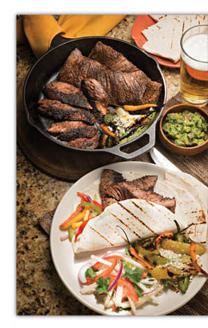 Black, cast iron skillet with cooked beef, small wooden container of guacamole, plate of grilled steak fajitas with sliced bell peppers and onions