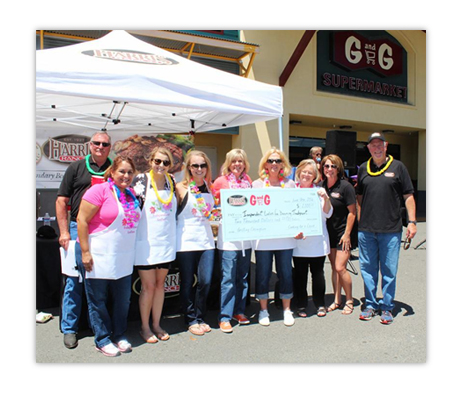 group of 8 men and women in aprons under a Harris Ranch tent canopy in parking lot in front of G G Market holding a large check signifying donation. Amount of check written too small to discern