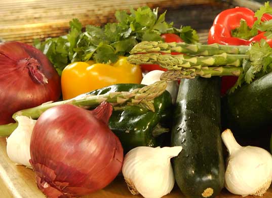 Close up of wood table top with fresh, raw veggies: red and white onions, asparagus, red and yellow bell peppers, cucumber