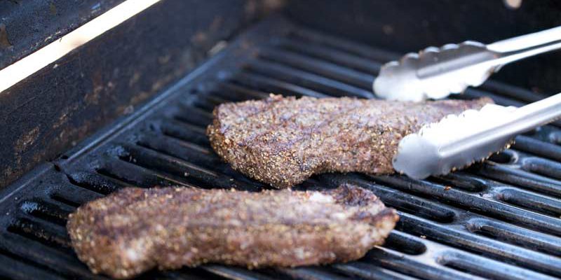 Close up of 3 NY steaks on grill. Steaks look ready to come off, have criss cross grill marks. Tongs in shot reaching for steak farthest from the camera