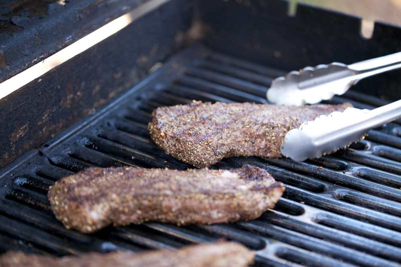 Close up of 3 NY steaks on grill. Steaks look ready to come off, have criss cross grill marks. Tongs in shot reaching for steak farthest from the camera