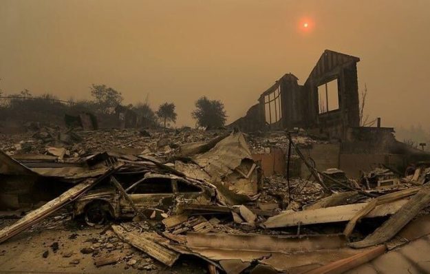 Kent Porter 1_preview Devastation from scene of recent fire. Burned out car, collapsed structure with only one partial wall remaining, yellow sun high in smokey sky, shrouded with orange ring