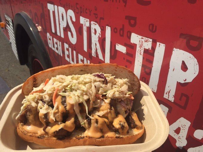 Grilled tri tip sandwich smothered with coleslaw and cheddar cheese, in paper take-out container, with Tips Tri-Tip lettering in white on red trolley in background