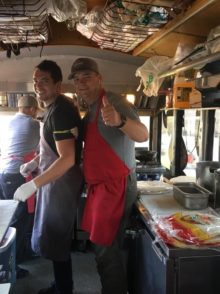 Two smiling men in BBQ aprons, one with thumbs up, inside catering trolley