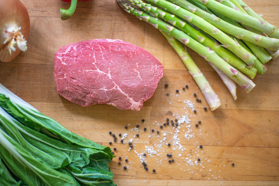 uncooked steak on cutting board with seasonings and uncooked vegetables